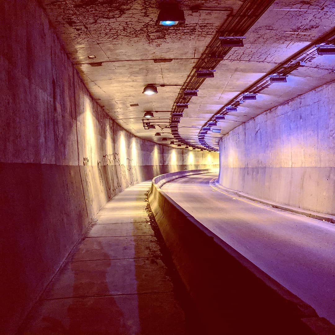 Empty tunnel with concrete walls and ceiling, illuminated by row of overhead lights, with a curved road passing through.