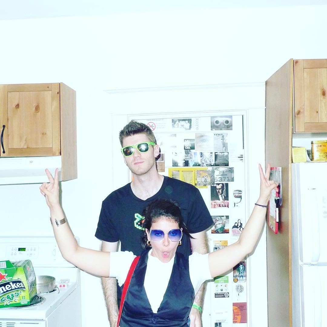 A woman with dark curly hair and blue sunglasses making a peace sign with both hands in front of a man with short dark hair and green sunglasses, in a kitchen with wooden cabinets and a refrigerator.
