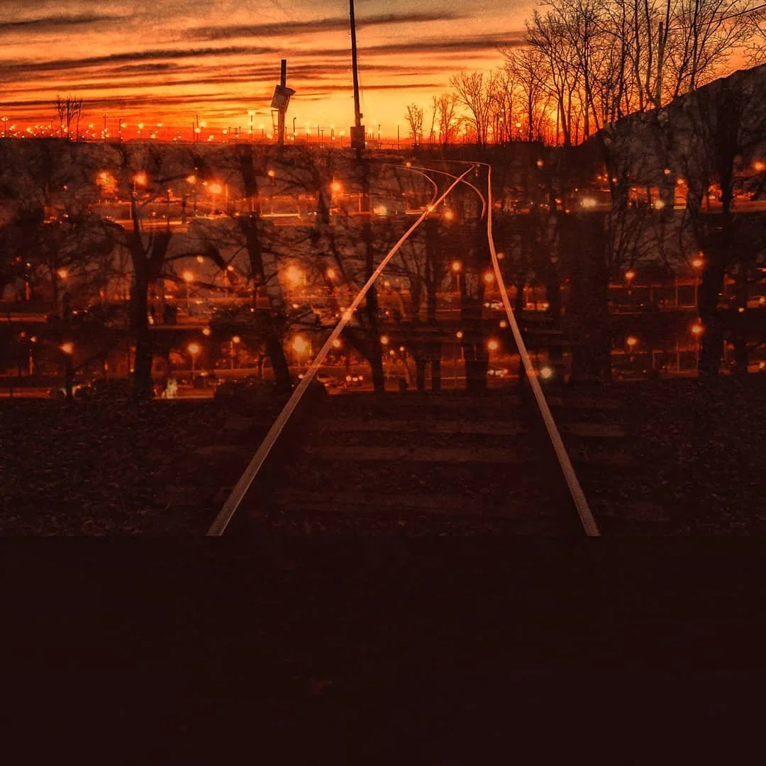 A railroad track at sunset, with a reflection of a city skyline with lights and leafless trees in the water.