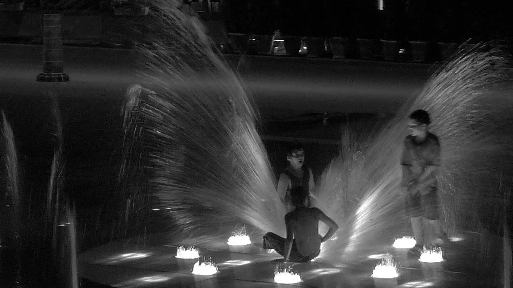 Children playing around small water fountains at night, with water spraying in arcs, in a dimly lit outdoor area.