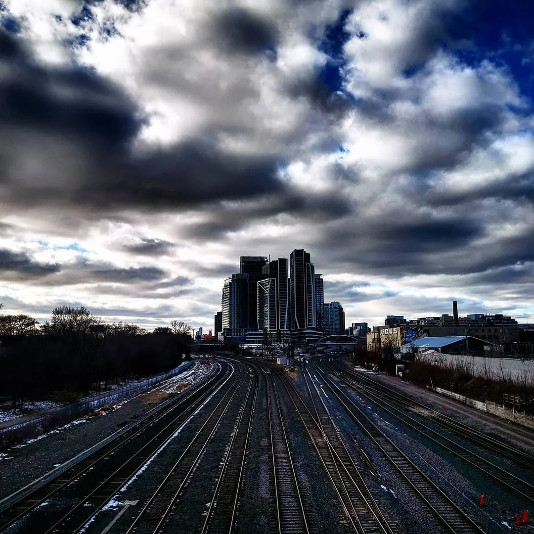 City skyline with tall modern buildings under cloudy sky, train tracks in foreground, some snow on ground.