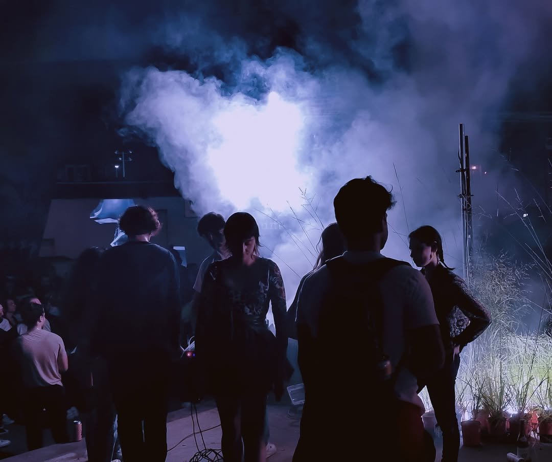 People gathered outdoors at night, illuminated by bright lights and surrounded by smoke or fog, with sparks flying in the background, possibly during a celebration or fireworks display.