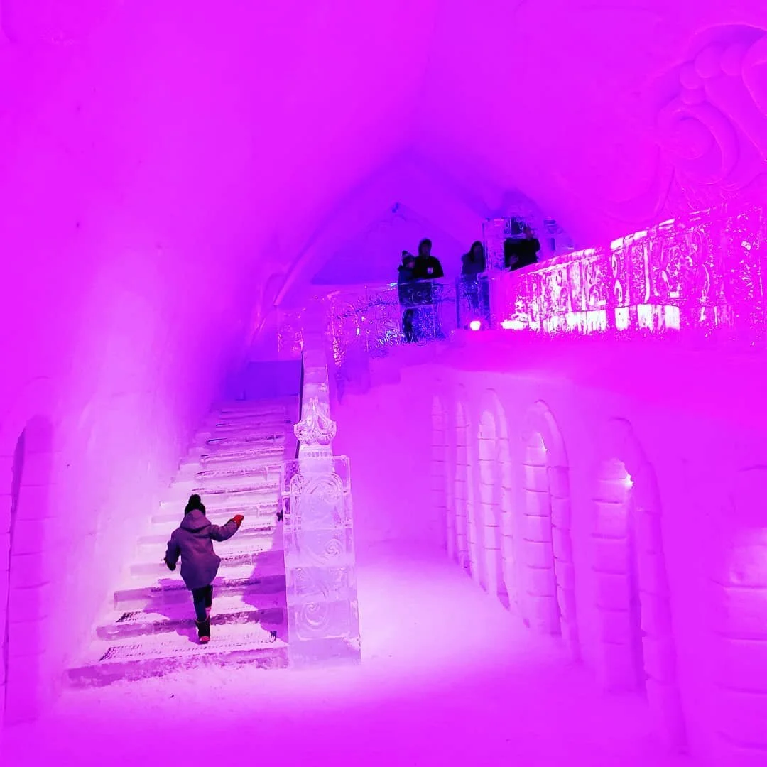 Ice sculpture hall illuminated with pink and purple lighting, featuring a staircase with carved ice railings and a child walking up the stairs, with three people standing on the ice balcony in the background.