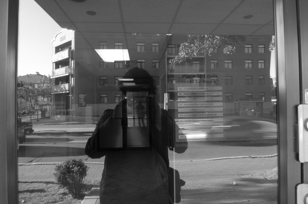 Reflection of a person taking a photo with a phone in a glass door, showing a street scene with buildings, cars, and a sidewalk outside.