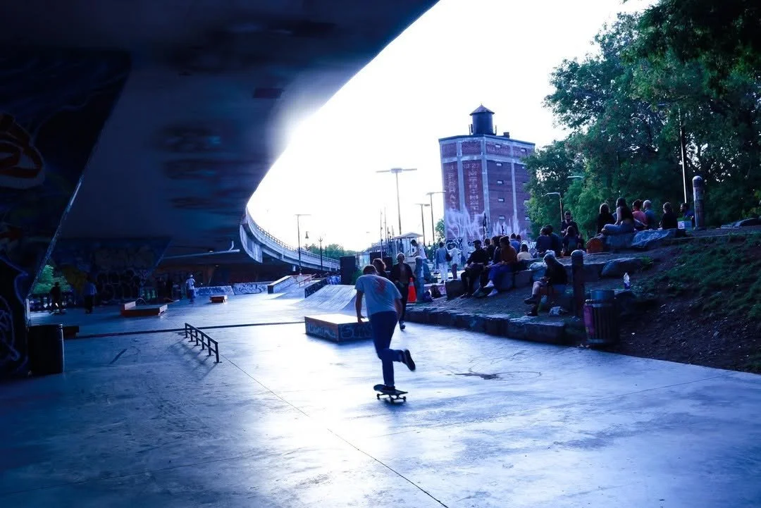Skateboarder skateboarding at an outdoor skatepark with a crowd sitting on rocks and green trees, urban buildings, and a bridge overhead.