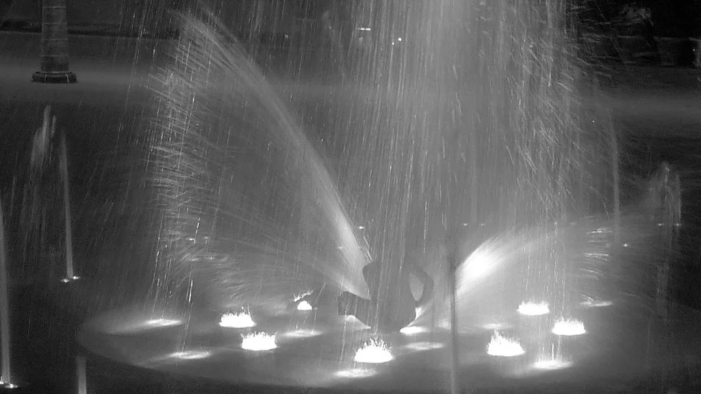 Nighttime scene of a person playing in a water fountain, with water jets illuminated by lights, creating a spray pattern.