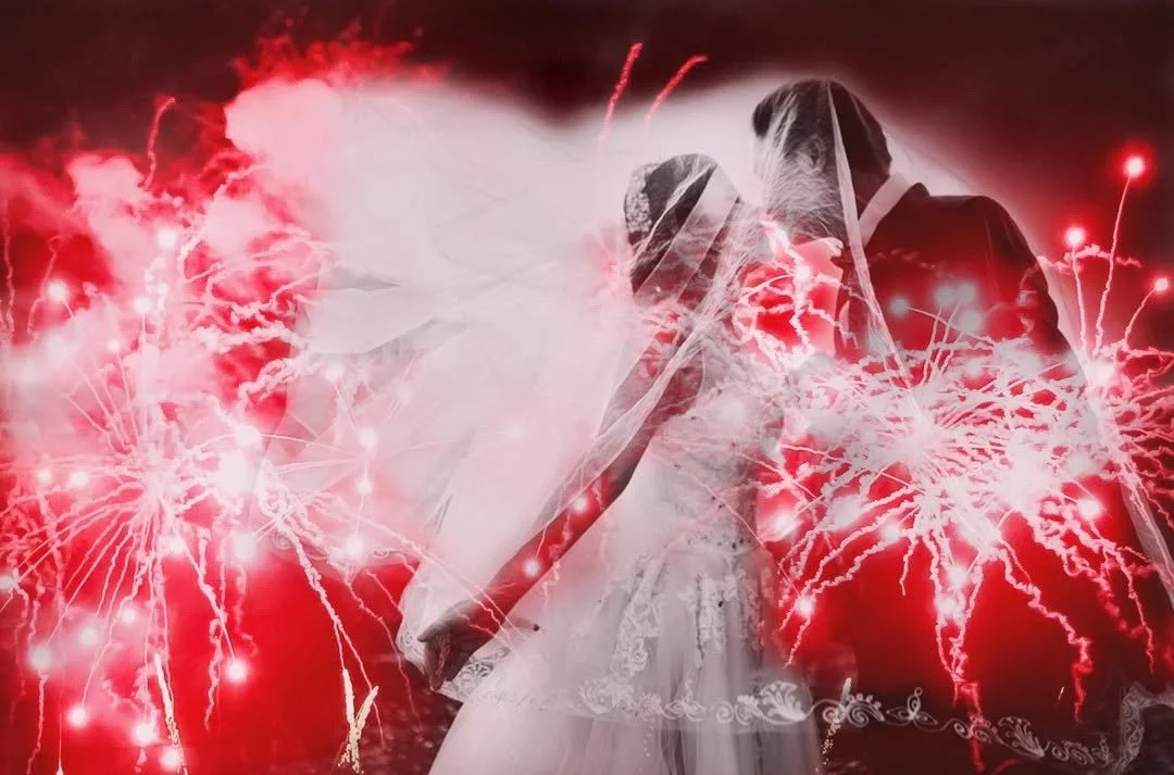 Couple dressed as bride and groom holding hands with red fireworks in background.