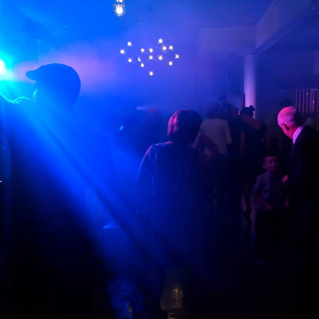 People dancing and socializing in a dark, blue-lit nightclub or party setting with a chandelier on the ceiling.