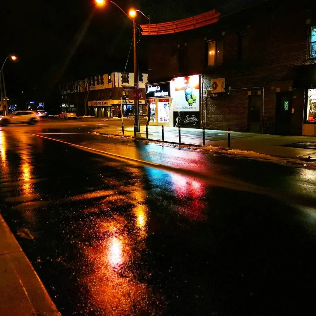 Nighttime city street scene with wet pavement reflecting streetlights and store signs. A stop sign, pole barriers, and storefronts including dental care and pharmacy are visible.