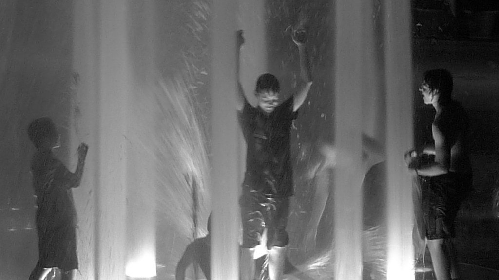 Three children playing with a water fountain at night, one in the middle jumping with arms up, another on the right holding a small object, and the third on the left watching.