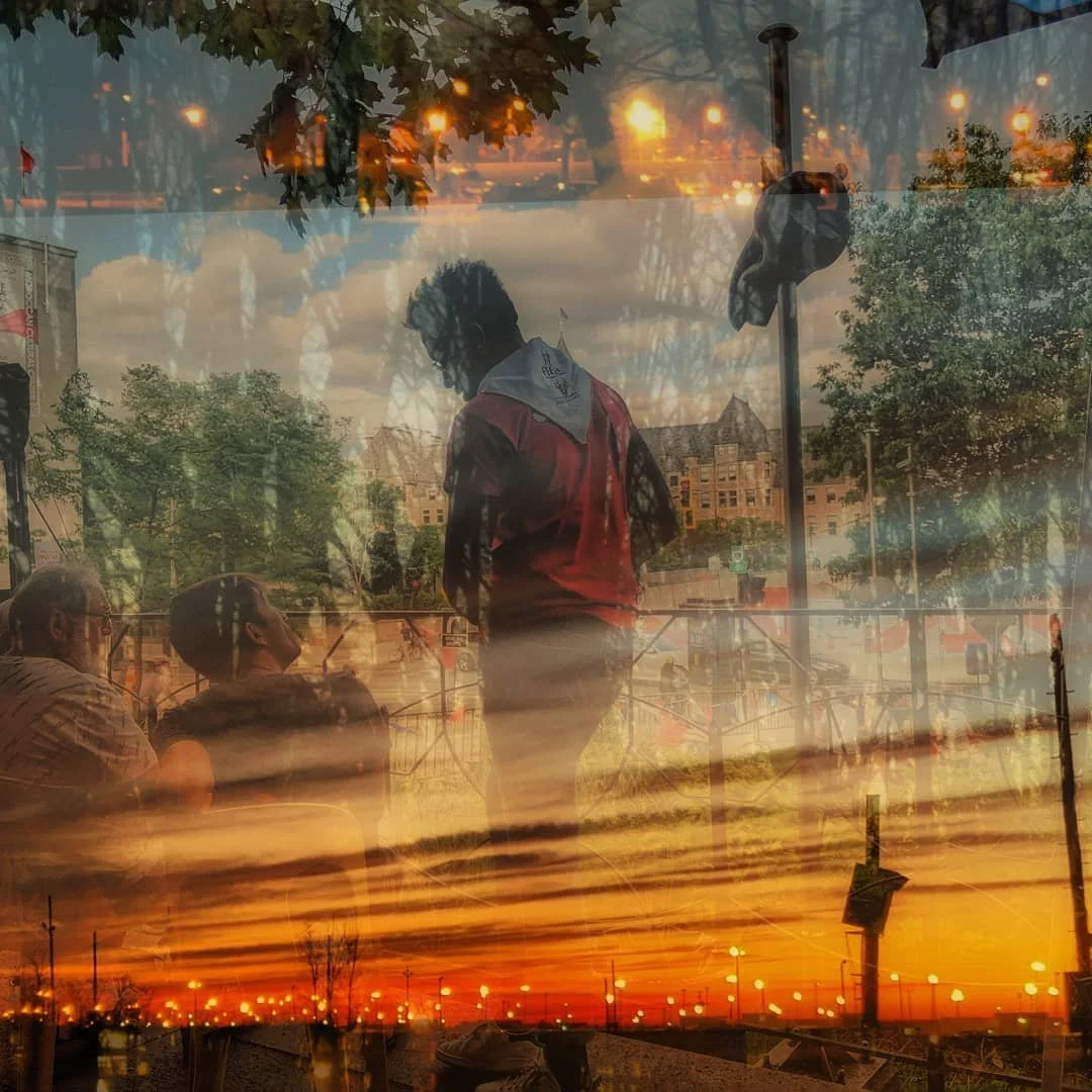 A layered photo with a sunset sky, cityscape, and outdoor scene with people, trees, and a person in a red shirt inside reflected through a glass window.