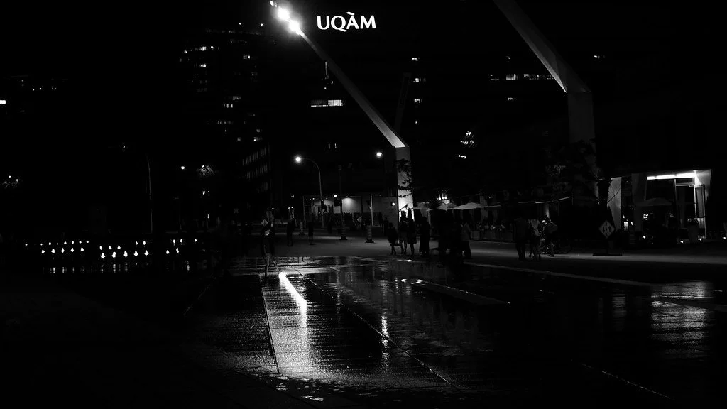 Night scene at a modern urban area with people walking on a wet street, illuminated by streetlights, with a building featuring the UQAM logo visible in the background.