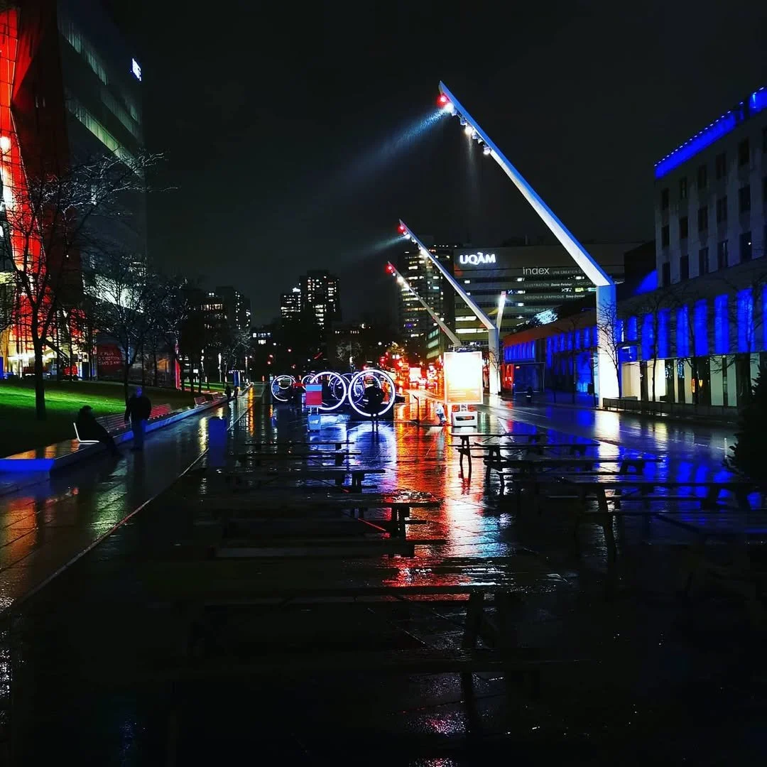 Nighttime view of a city street with illuminated buildings, colorful neon lights, wet pavement reflecting the lights, benches, and a few people walking.