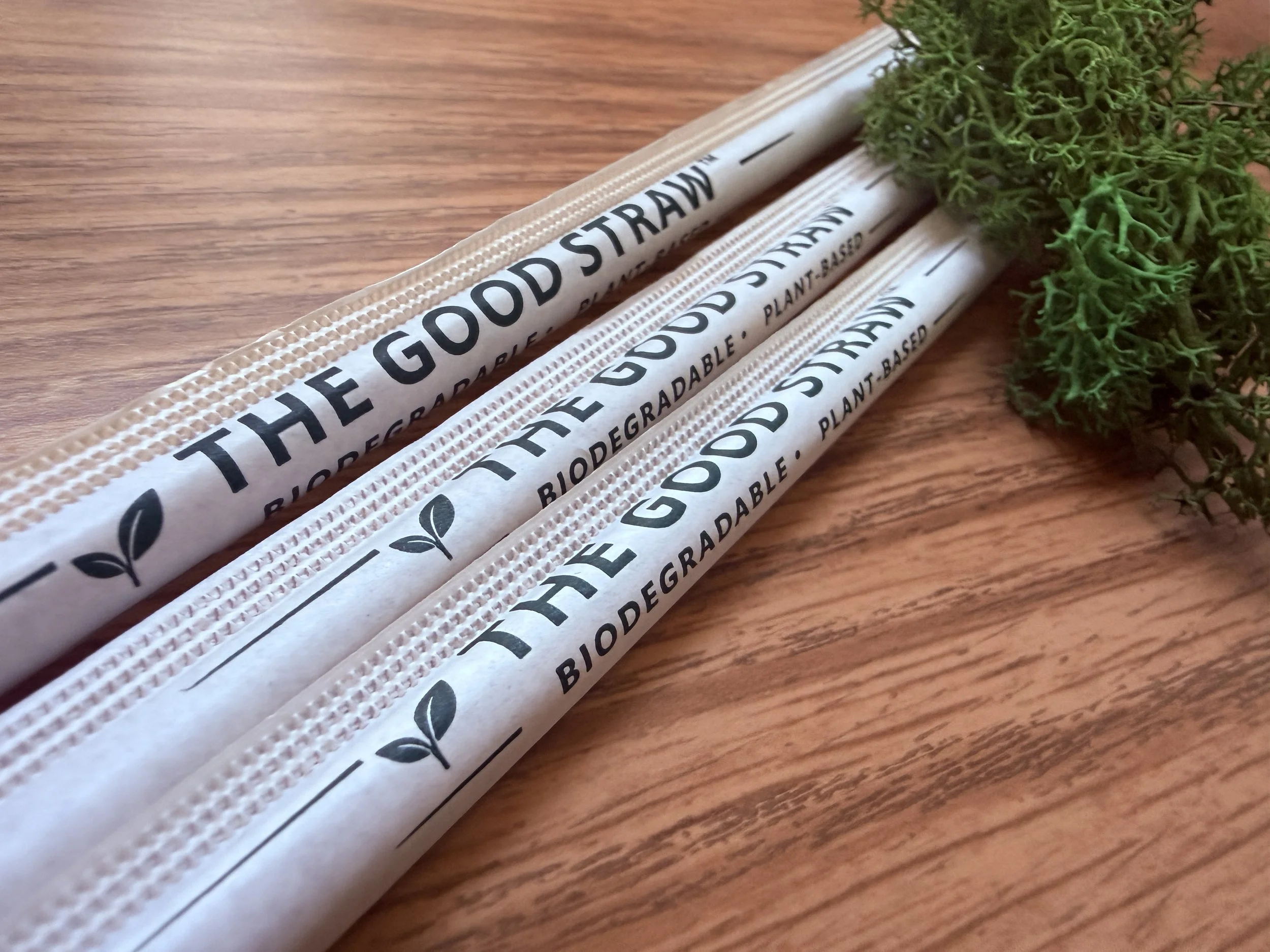 Three biodegradable paper straws with black text reading 'THE GOOD STRAW' and a small leaf graphic, next to a green moss or lichen on a wooden surface.