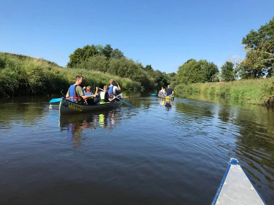 People canoeing on a calm river under clear blue skies, surrounded by green trees and bushes.