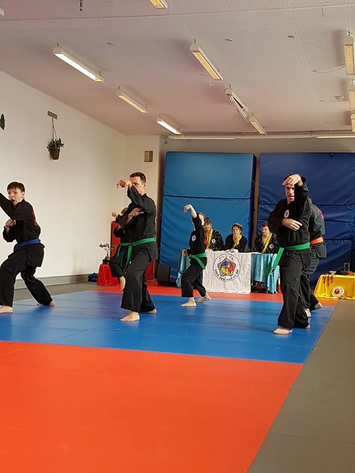Martial arts demonstration with four students performing moves on the mat in an indoor dojo, with a panel of judges sitting behind a table in the background.