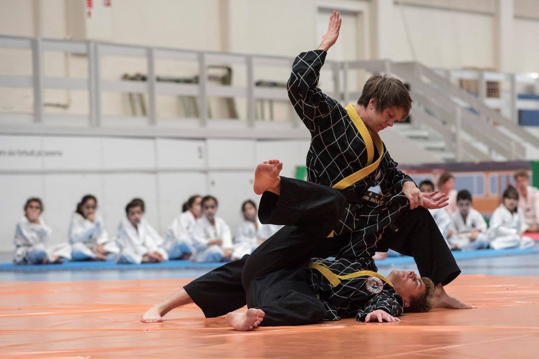 Two martial artists practicing Brazilian Jiu-Jitsu on an orange mat, with spectators in martial arts uniforms watching in the background.