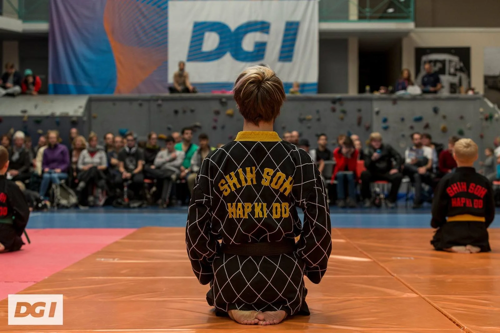 A young martial artist kneeling on a mat in a martial arts competition, wearing a black gi with yellow patches, facing an audience seated in front of a large banner with the letters 'DGI', and a rock climbing wall in the background.