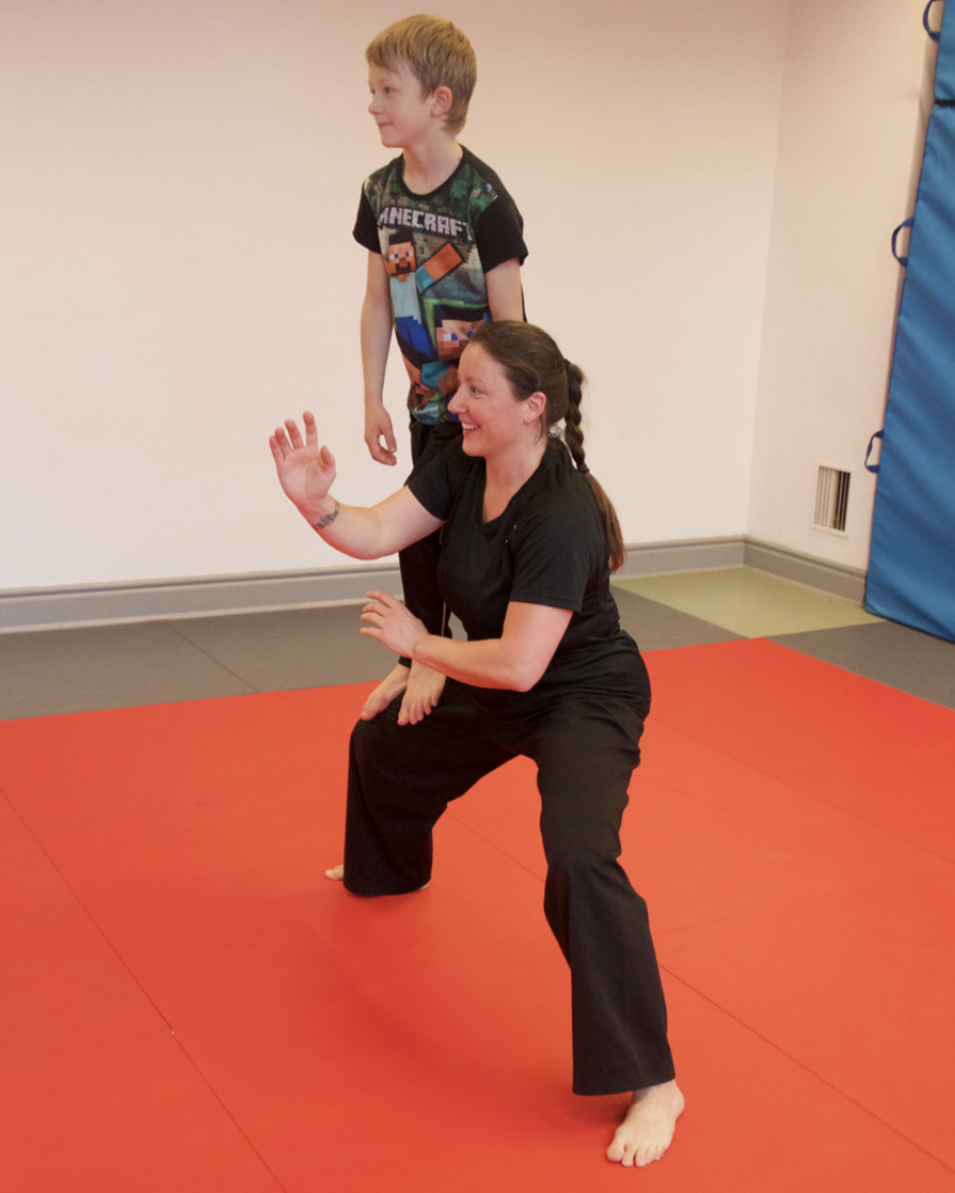 A woman practicing martial arts on a red mat with a young boy sitting on her back, in a room with beige walls and blue padding on the side.