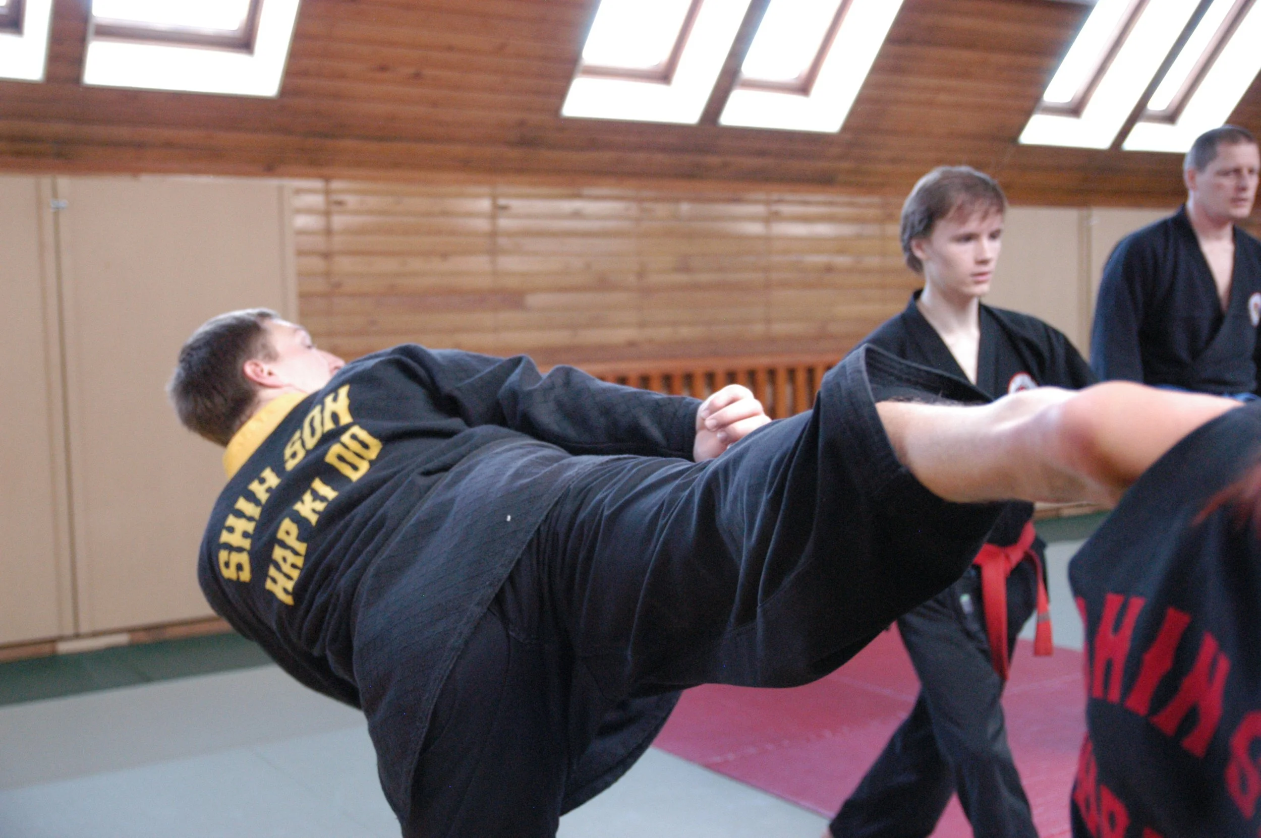 A person practicing martial arts in a training dojo, wearing a black gi with yellow lettering on the back, executing a high kick while others in black gis observe.
