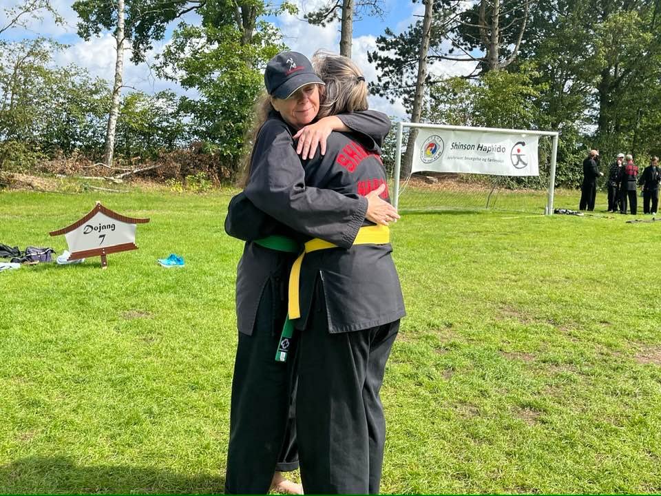 Two women in martial arts uniforms hugging each other outdoors on a grassy field, with trees and a banner in the background.