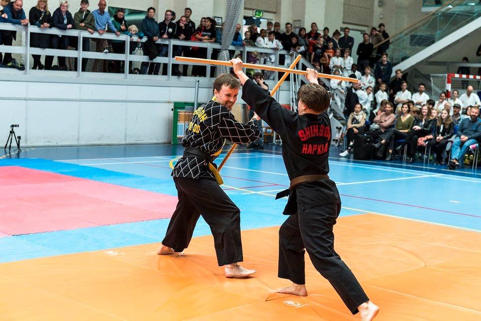 Two martial artists practicing martial arts with wooden sticks in front of an audience in a gymnasium, with spectators watching from the stands.