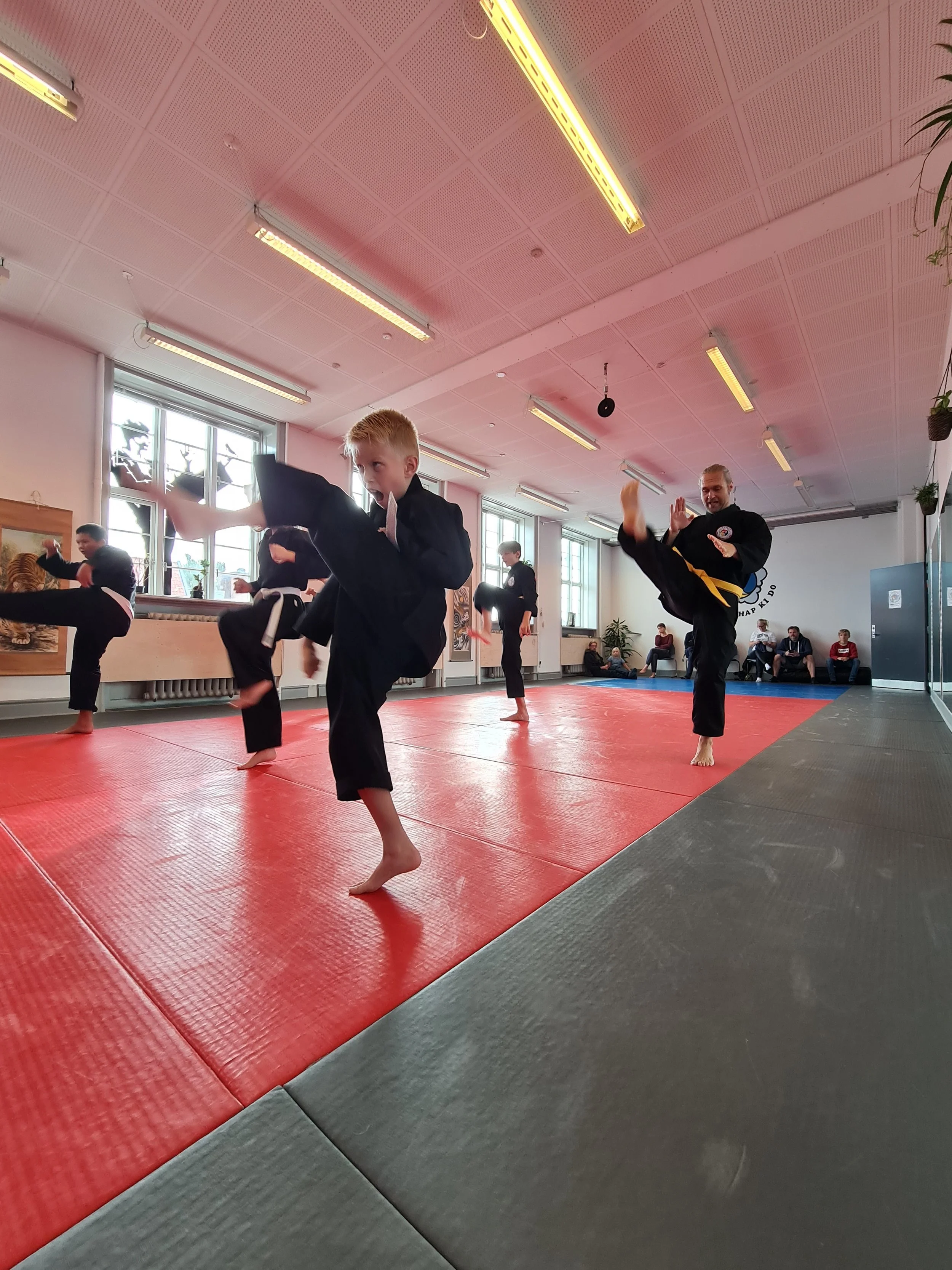 Kids in martial arts uniforms practicing kicks in a gym with mats, under fluorescent lighting, with spectators sitting in the background.