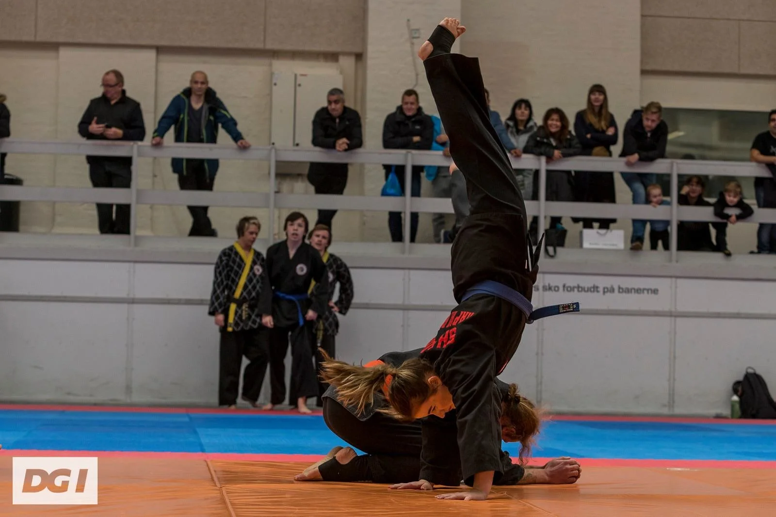 Two women practicing Brazilian Jiu-Jitsu on a mat, with one woman standing in a handstand position over the other on her hands and knees, while spectators watch from the background.