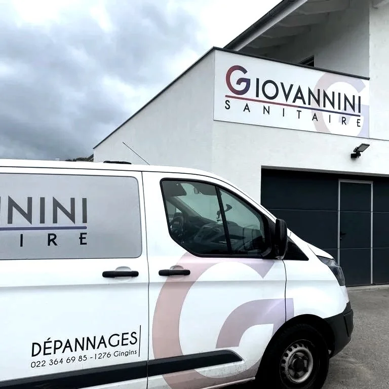 Une camionnette blanche avec logo et texte 'Giovanni Sanitaire, Dépannages, 022 364 69 85 - 1276 Gingin' garée devant un bâtiment avec un panneau indiquant le même nom, extérieur d'une entreprise de services sanitaires.