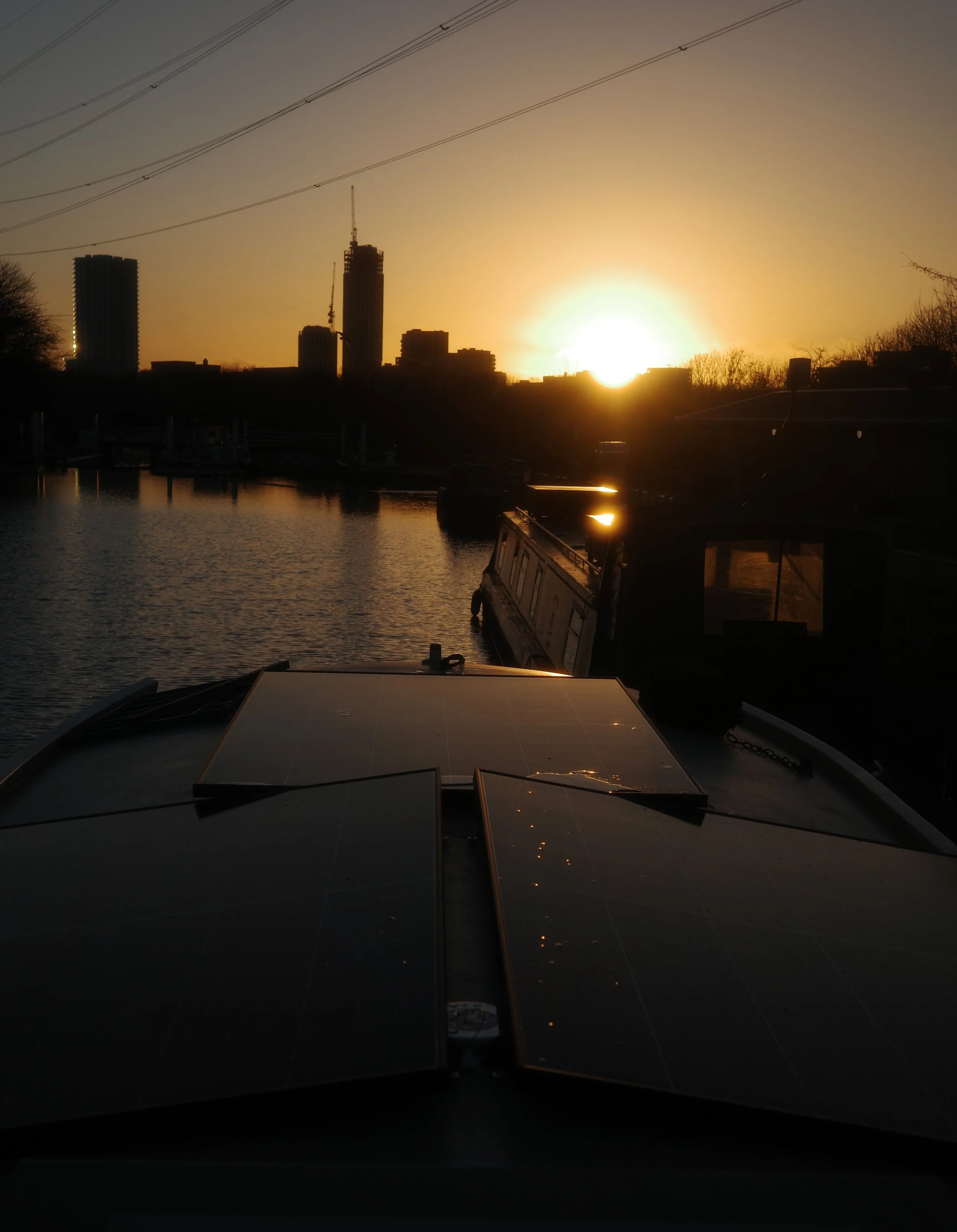 	
Sunset over a river with boats, city skyline with tall buildings, and power lines in the background. Taken whilst standing on top of the sauna boat