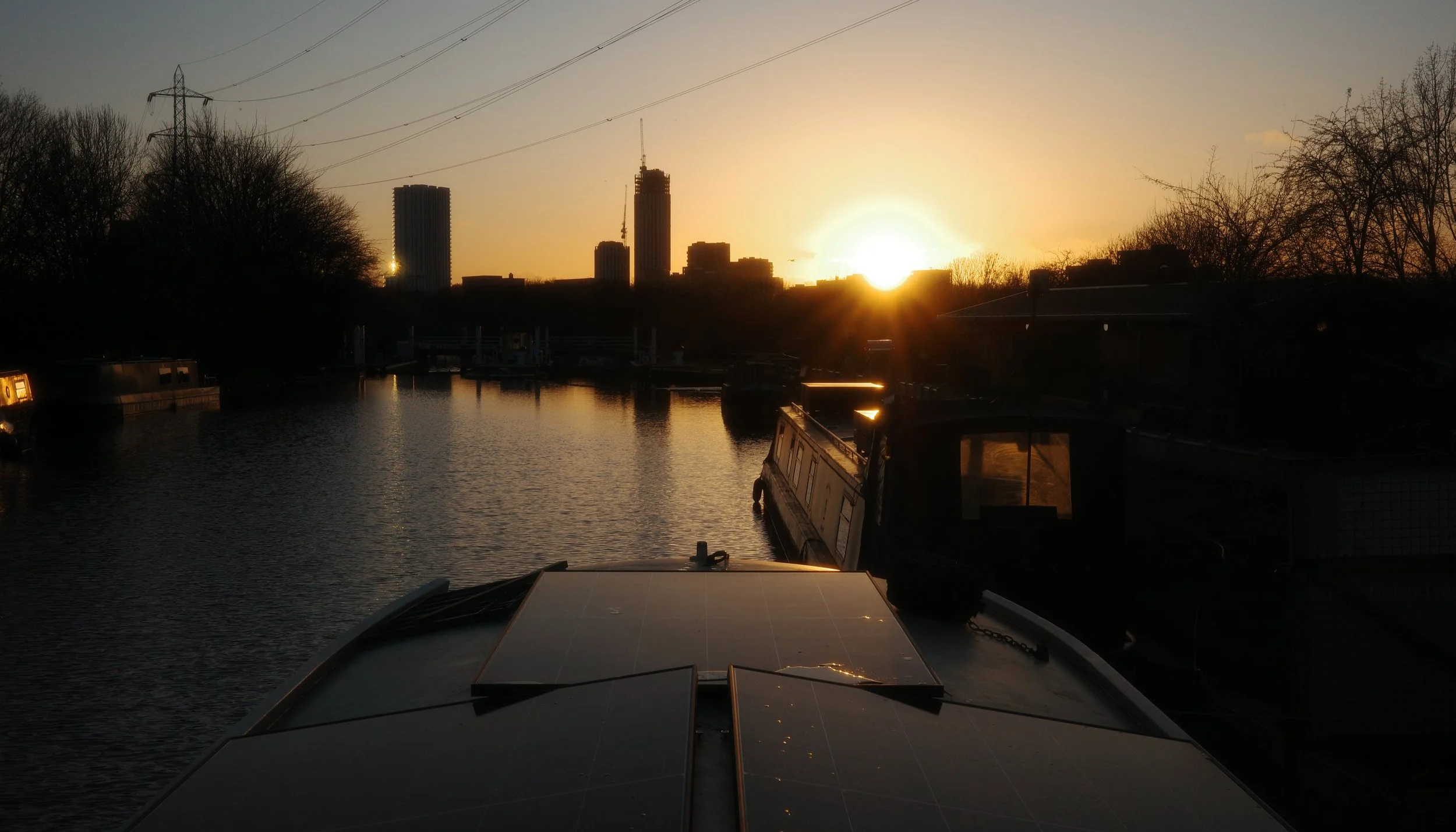Sunset over a canal with boats and tall buildings in the city skyline, trees on the sides, and solar panels on SOAK's Sauna boat in the foreground.