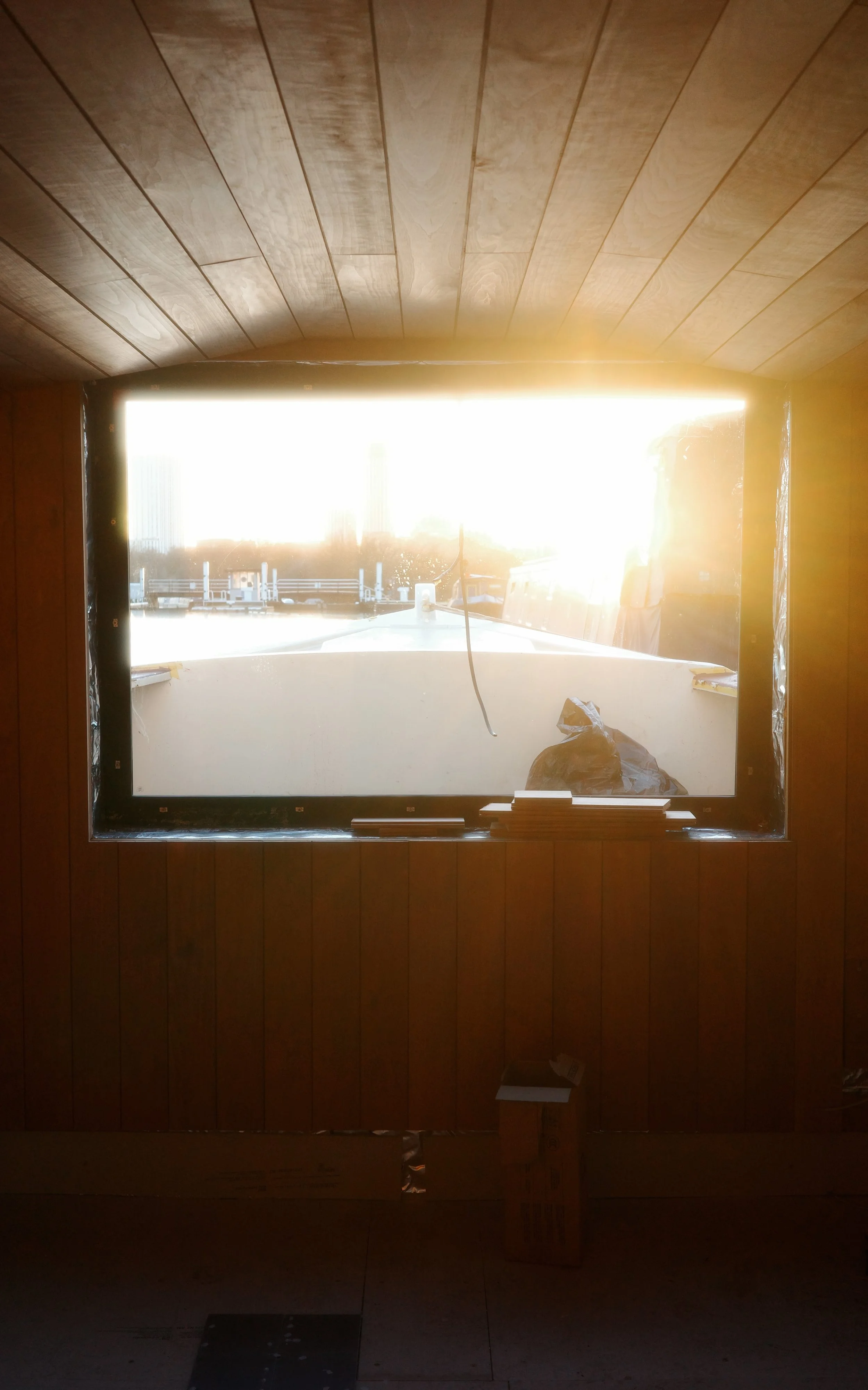 A construction site view through a rectangular sauna window with a wooden frame on the sauna boat, showing bright sunlight outside illuminating the scene, with construction materials and a partially built structure visible in the background.