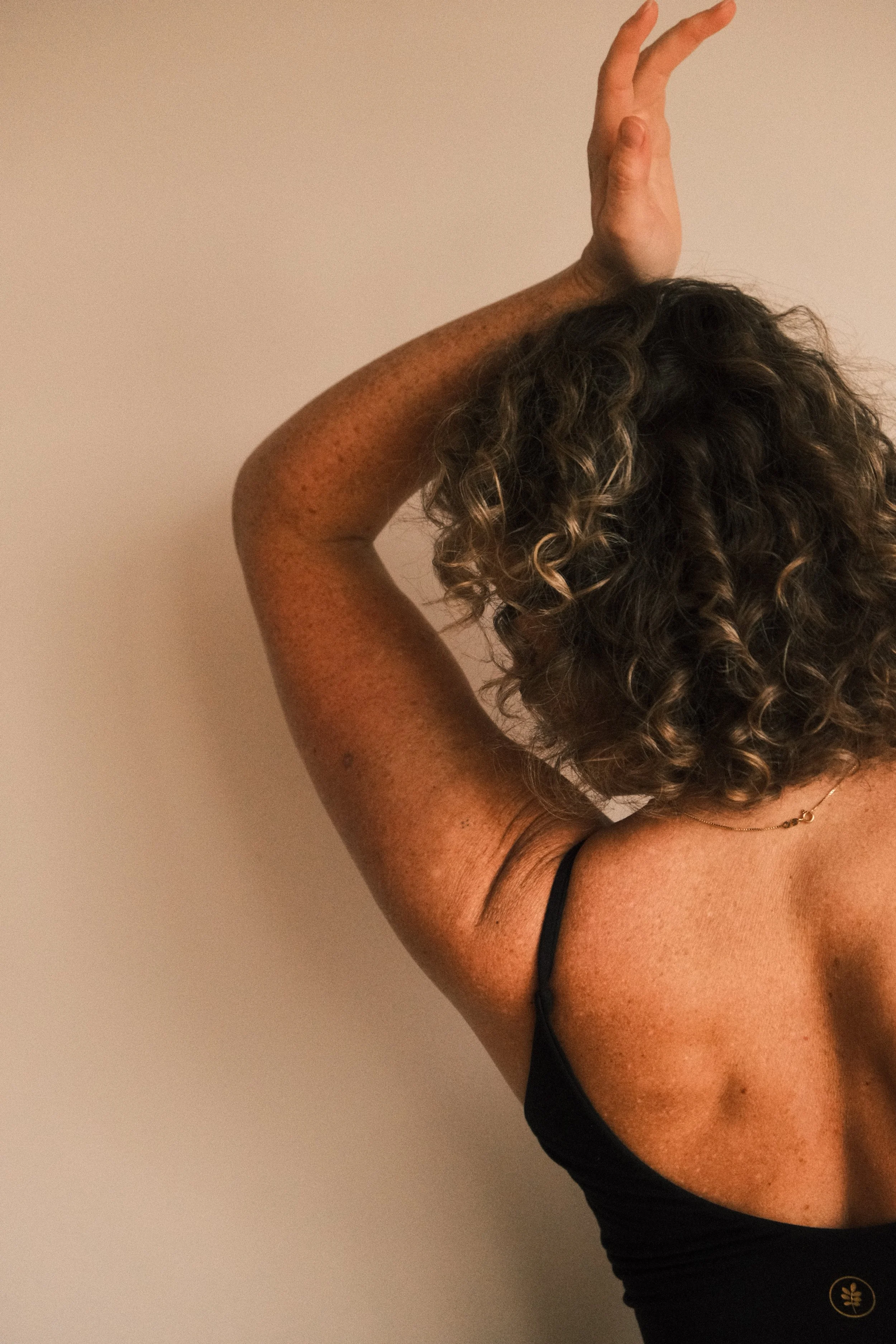 A person with curly hair and freckled skin wearing a black top and a gold necklace, posing with one arm raised and hand behind their head against a plain light-colored wall.