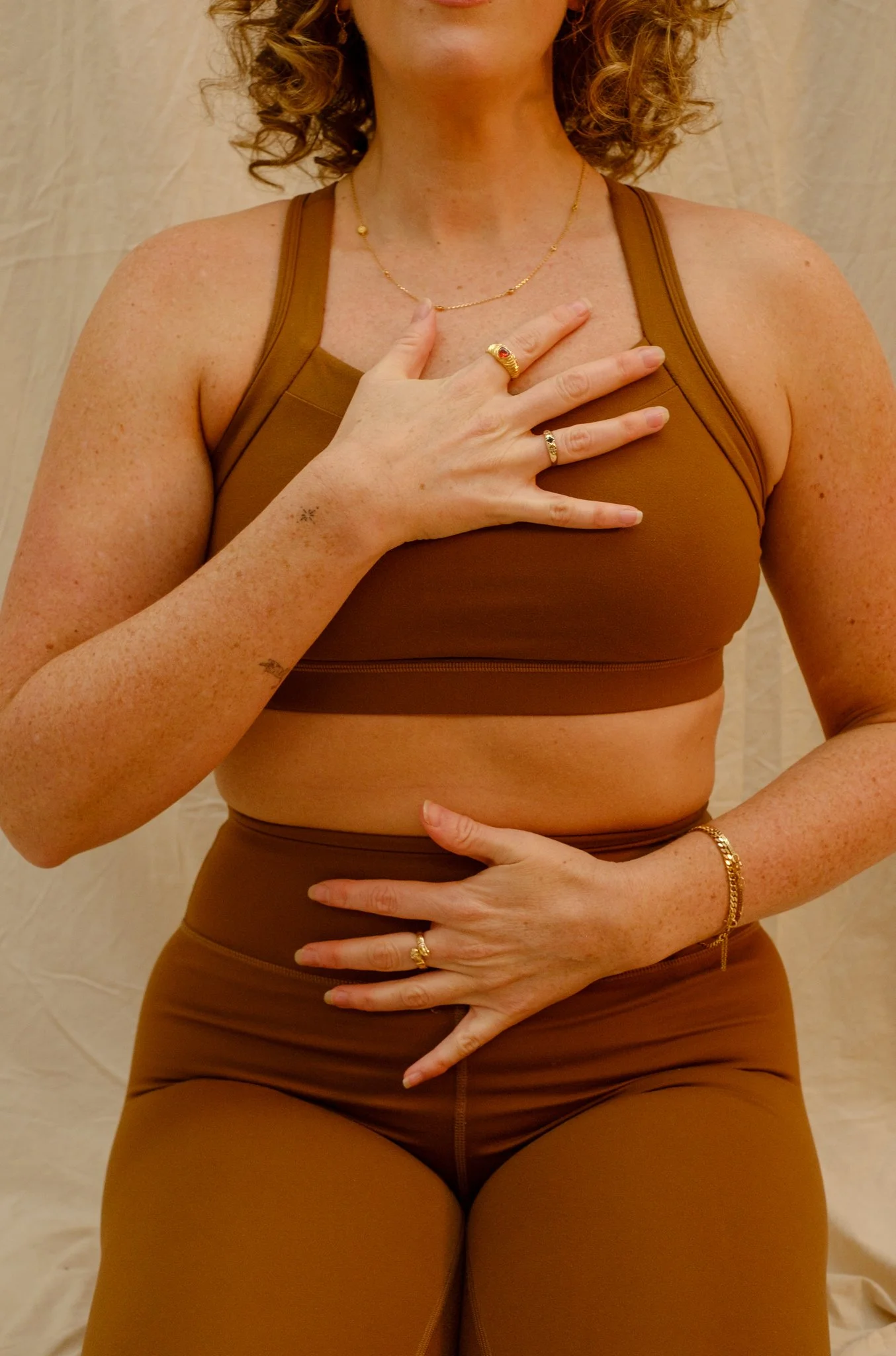 Woman wearing a brown sports bra and matching leggings, showing jewelry including rings, bracelets, a necklace, and her hand on her chest and stomach, with curly hair and light skin.