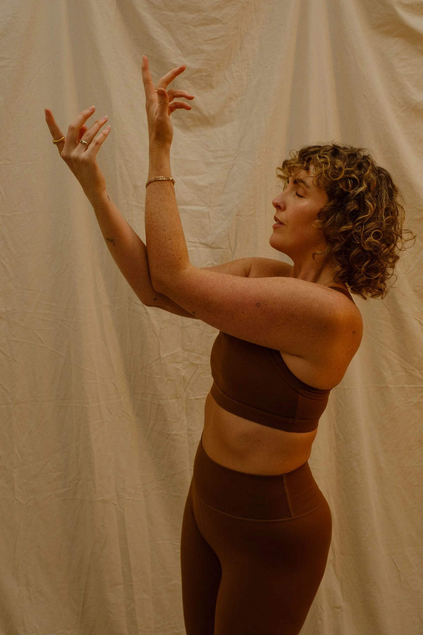 A woman with curly hair wearing a brown sports bra and matching leggings stands in profile against a neutral backdrop. Her eyes are closed, and her arms are raised with hands in expressive positions.