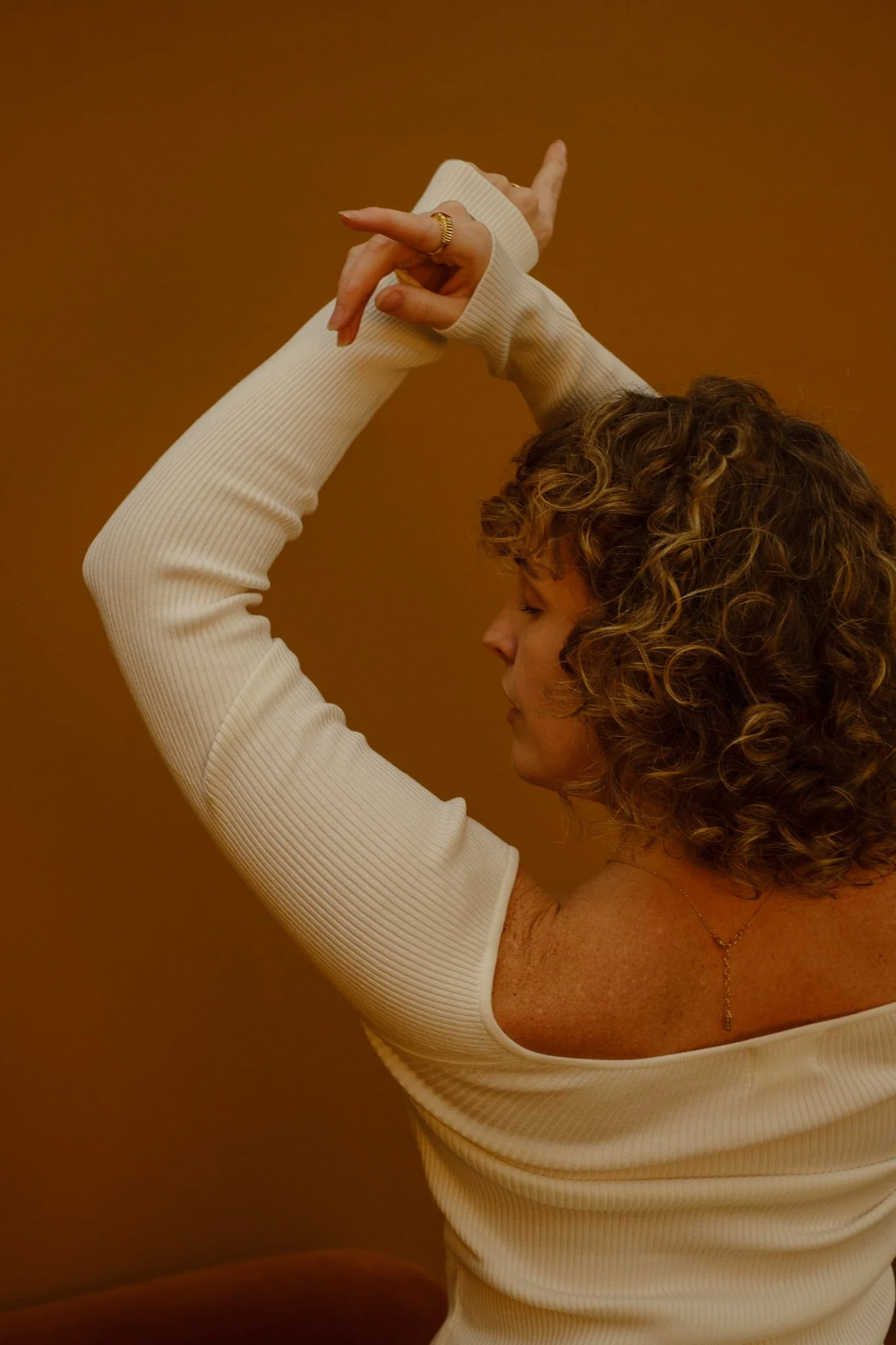 A woman with curly hair in a white ribbed long-sleeve top with her arms raised, crossing hands above her head against a brown background.