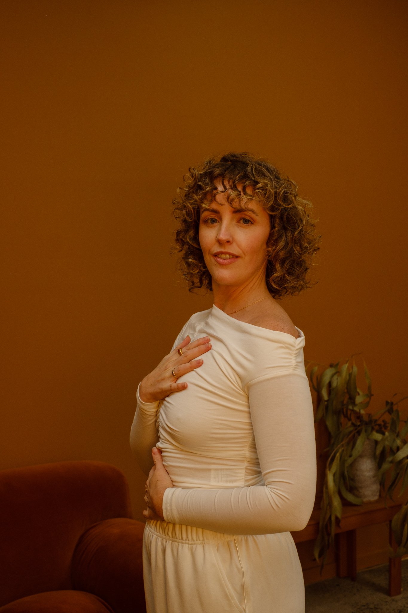 A woman with curly blonde hair wearing a cream long-sleeve dress standing in a room with brown walls and a plant in the background.