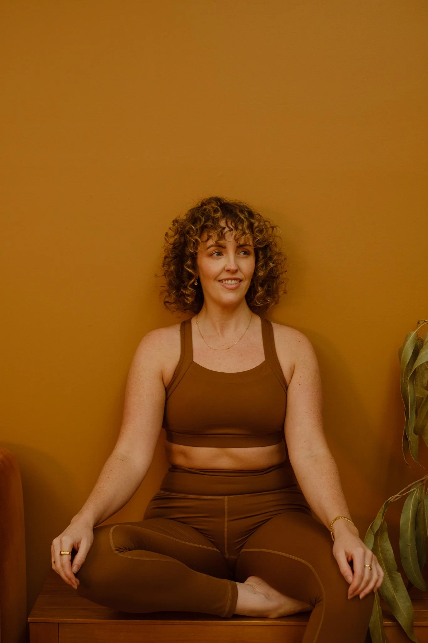 A woman with curly hair sitting cross-legged in a meditative pose against a mustard-colored wall, wearing brown athletic wear.