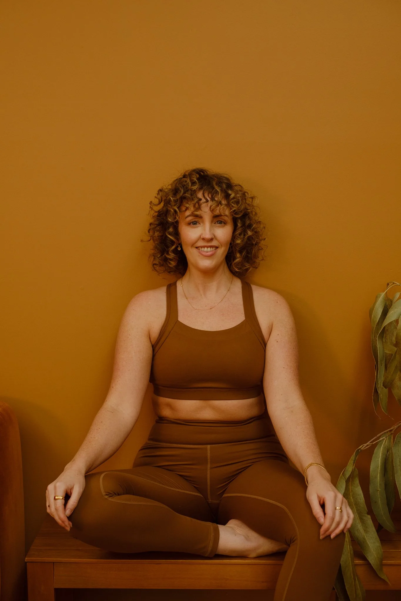 A woman with curly hair practicing yoga or meditation in a seated position, cross-legged on a wooden platform against a warm yellow wall, with a plant visible on the right side.