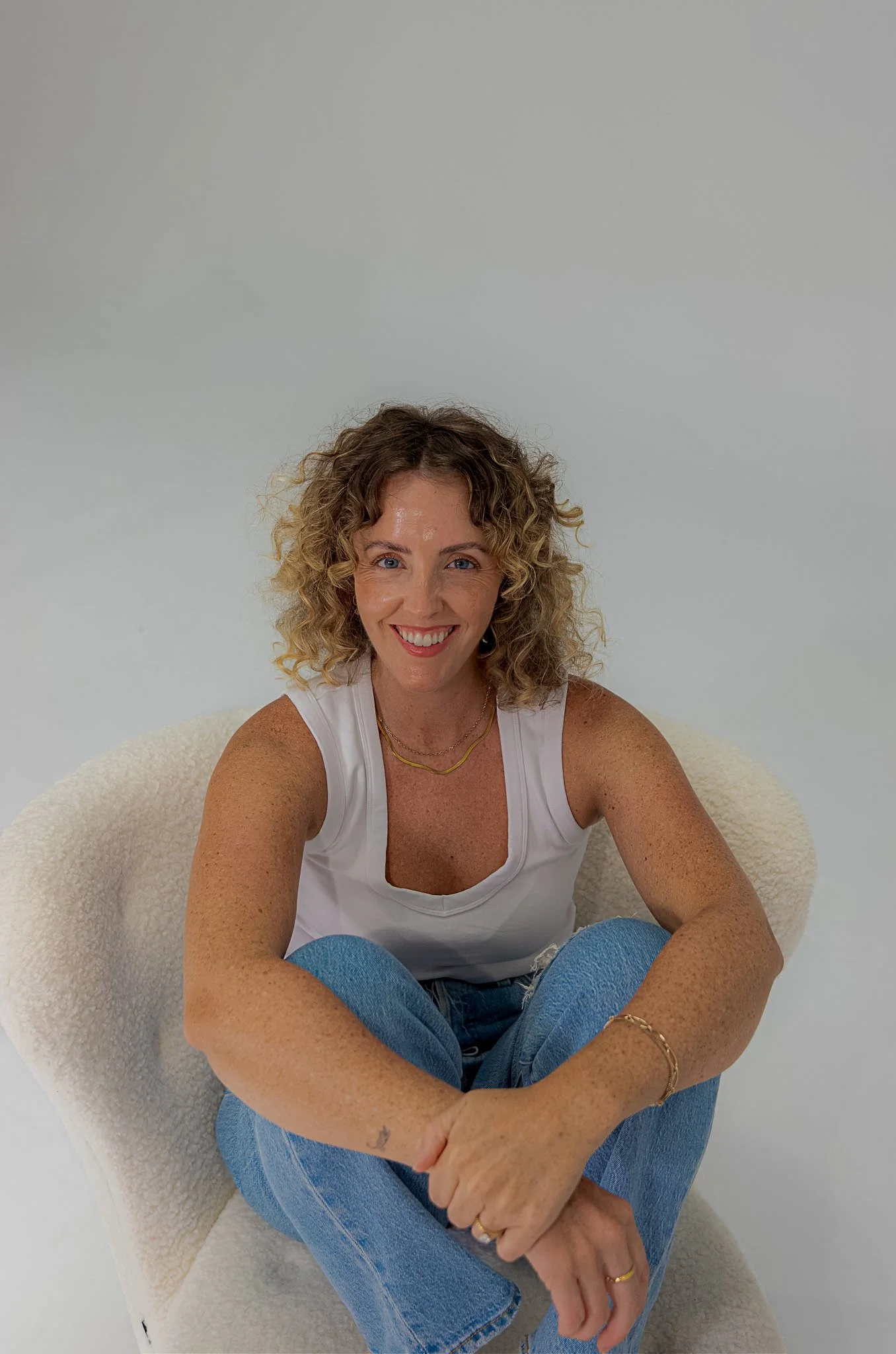 A woman with curly blonde hair sitting on a white, textured chair against a plain, light-colored wall. She is smiling, wearing a white tank top, blue jeans, and gold jewelry, including rings, a bracelet, and layered necklaces.