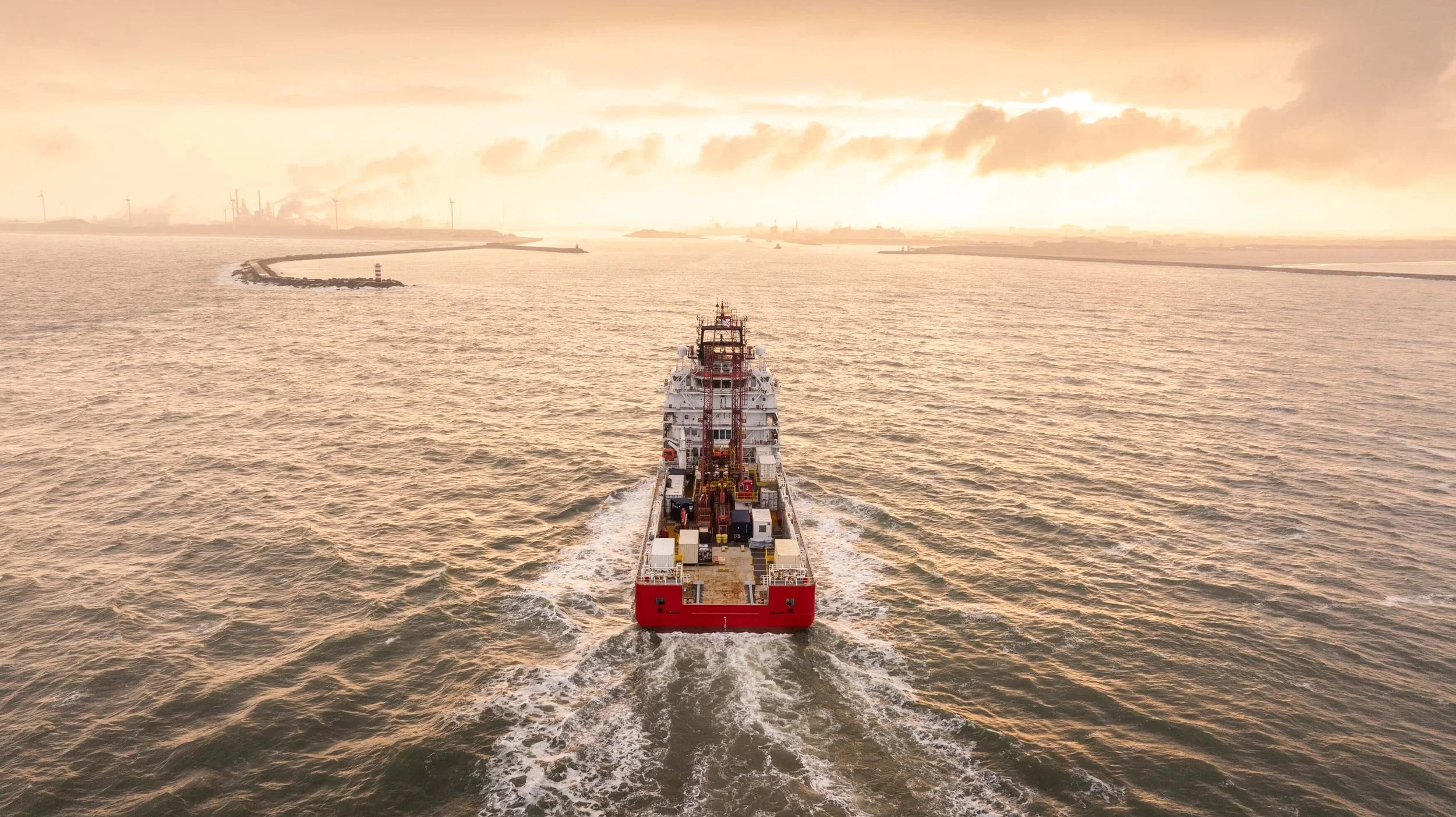 A cargo ship sailing in the open sea at sunset, with industrial structures and wind turbines in the distance.
