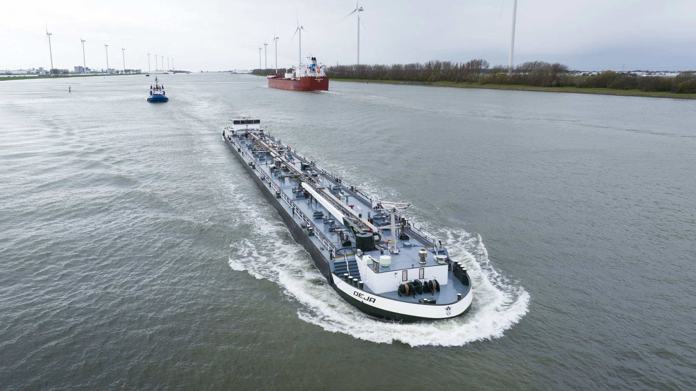A large cargo ship with the name 'OEJA' sailing in a wide waterway, accompanied by a smaller tugboat, with wind turbines and windmill blades visible in the distance.