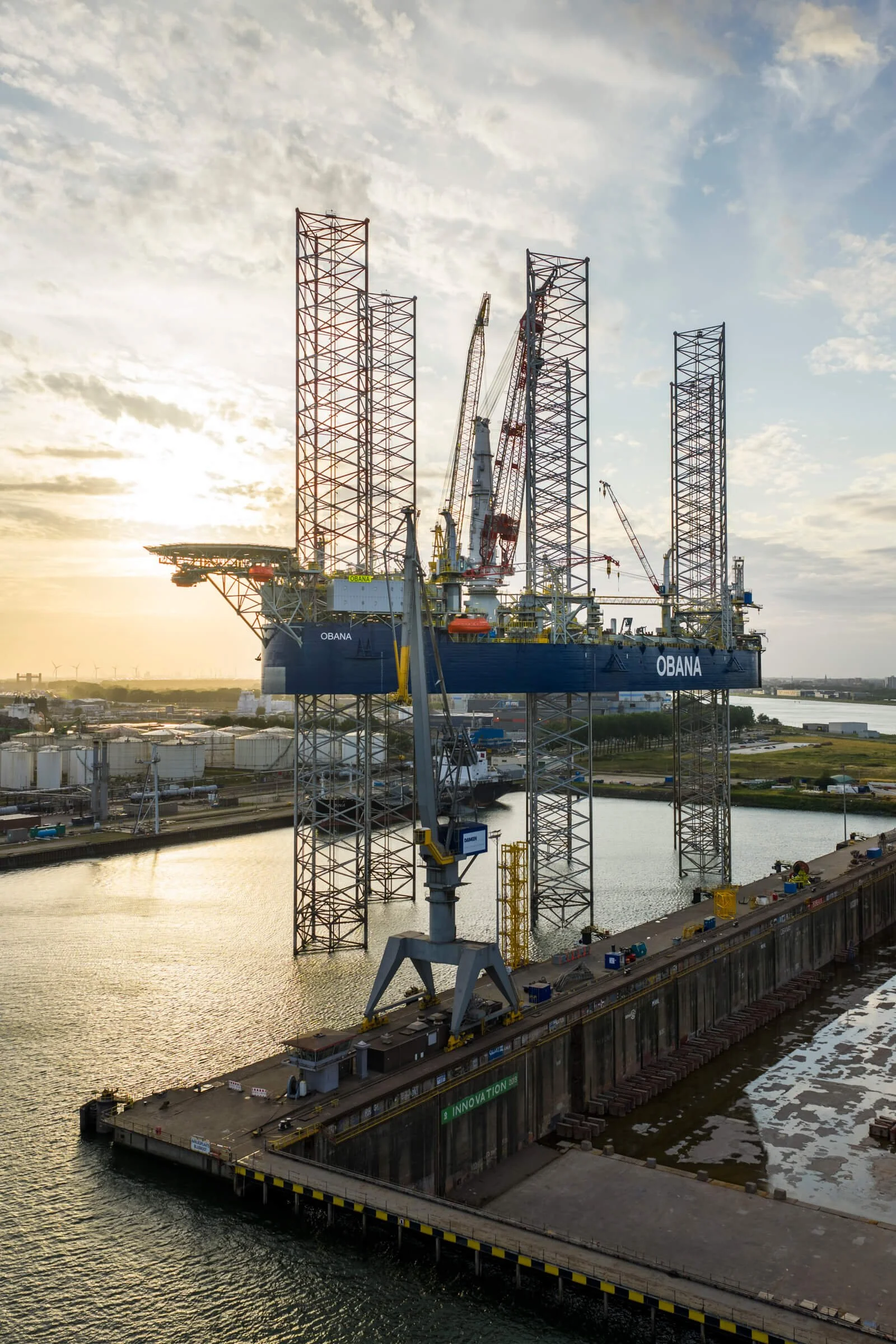 Offshore oil drilling platform named OBANA in the water at sunset, with cranes and structures on the platform and a cityscape in the background.