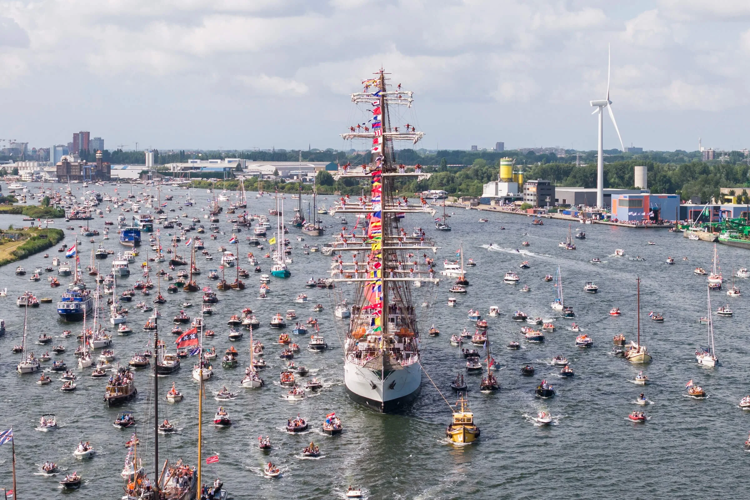A large ship decorated with colorful flags sailing through numerous smaller boats on a body of water, with a cityscape, wind turbine, and industrial buildings in the background.