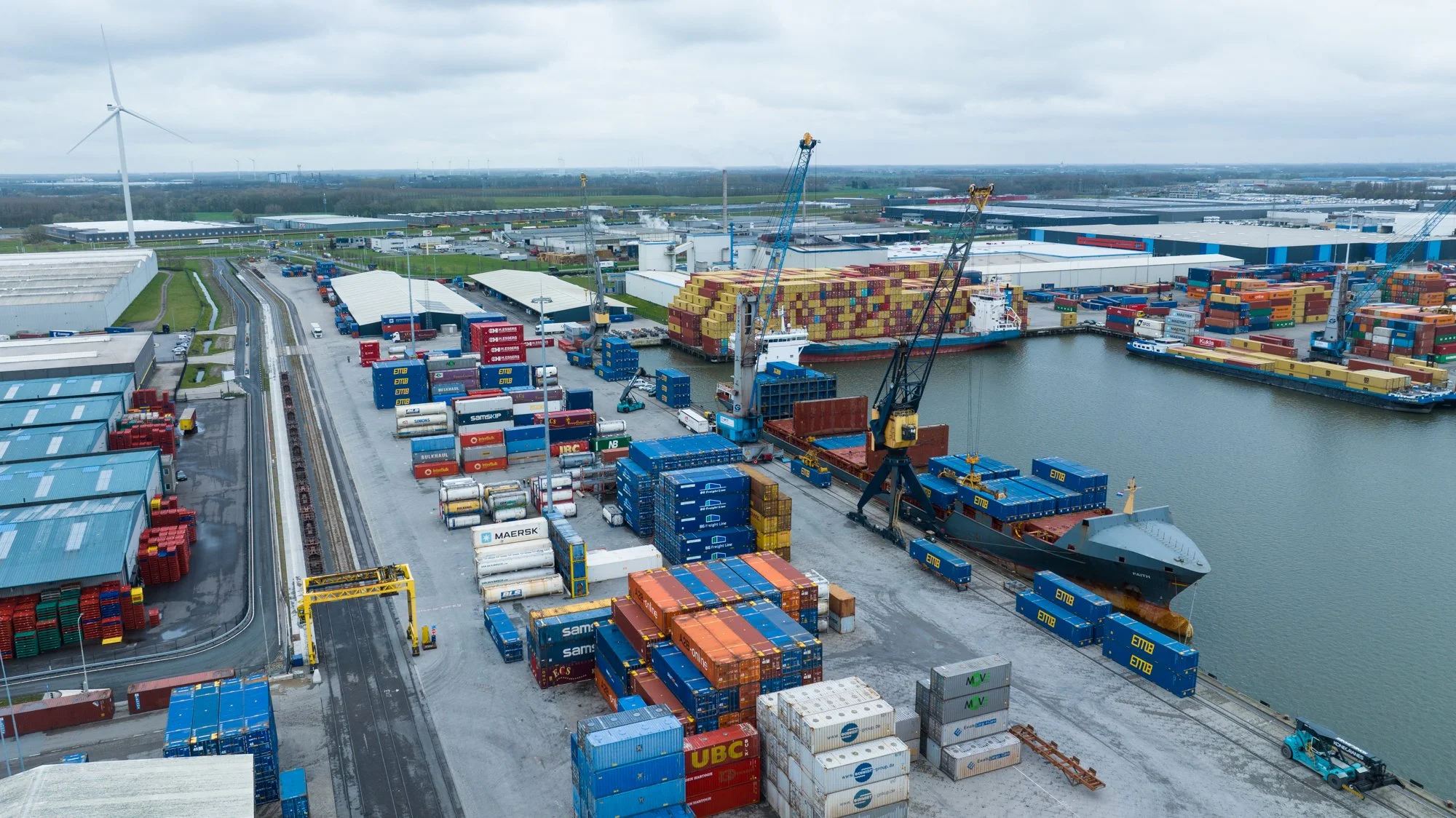 Aerial view of a busy container port with cargo ships, stacked shipping containers, cranes, and warehouses under an overcast sky.