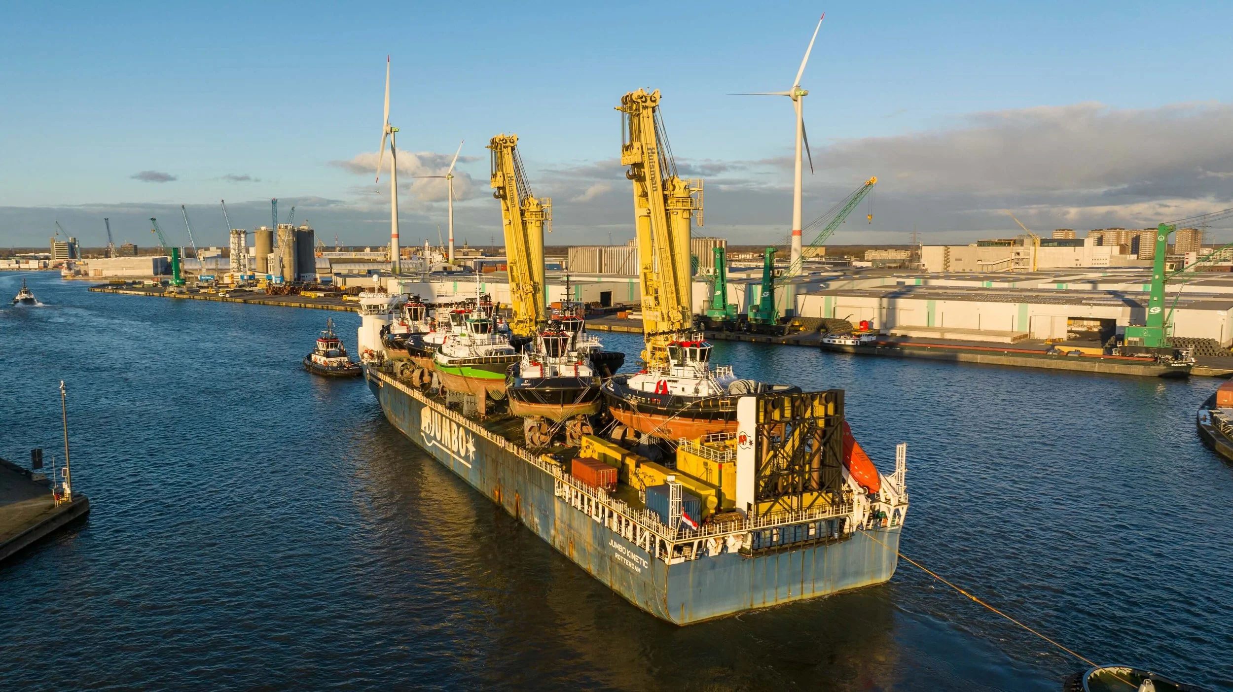 Cargo ship docked at a port with boats and cranes, wind turbines in the background, and a partly cloudy sky.