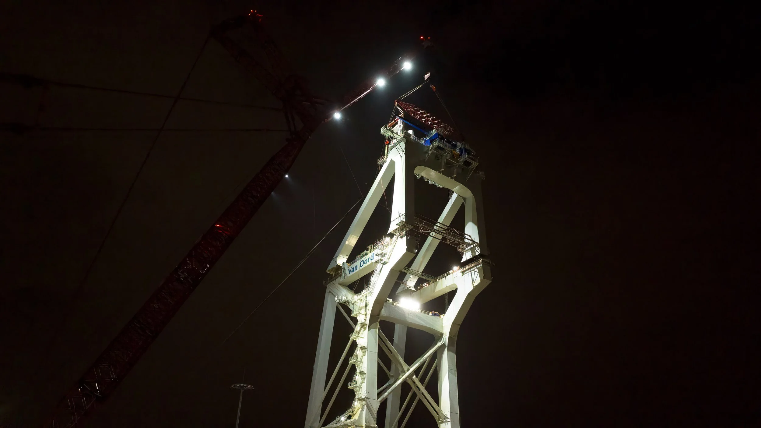 Nighttime view of a large white construction or industrial tower with a crane extending beside it, illuminated against the dark sky.