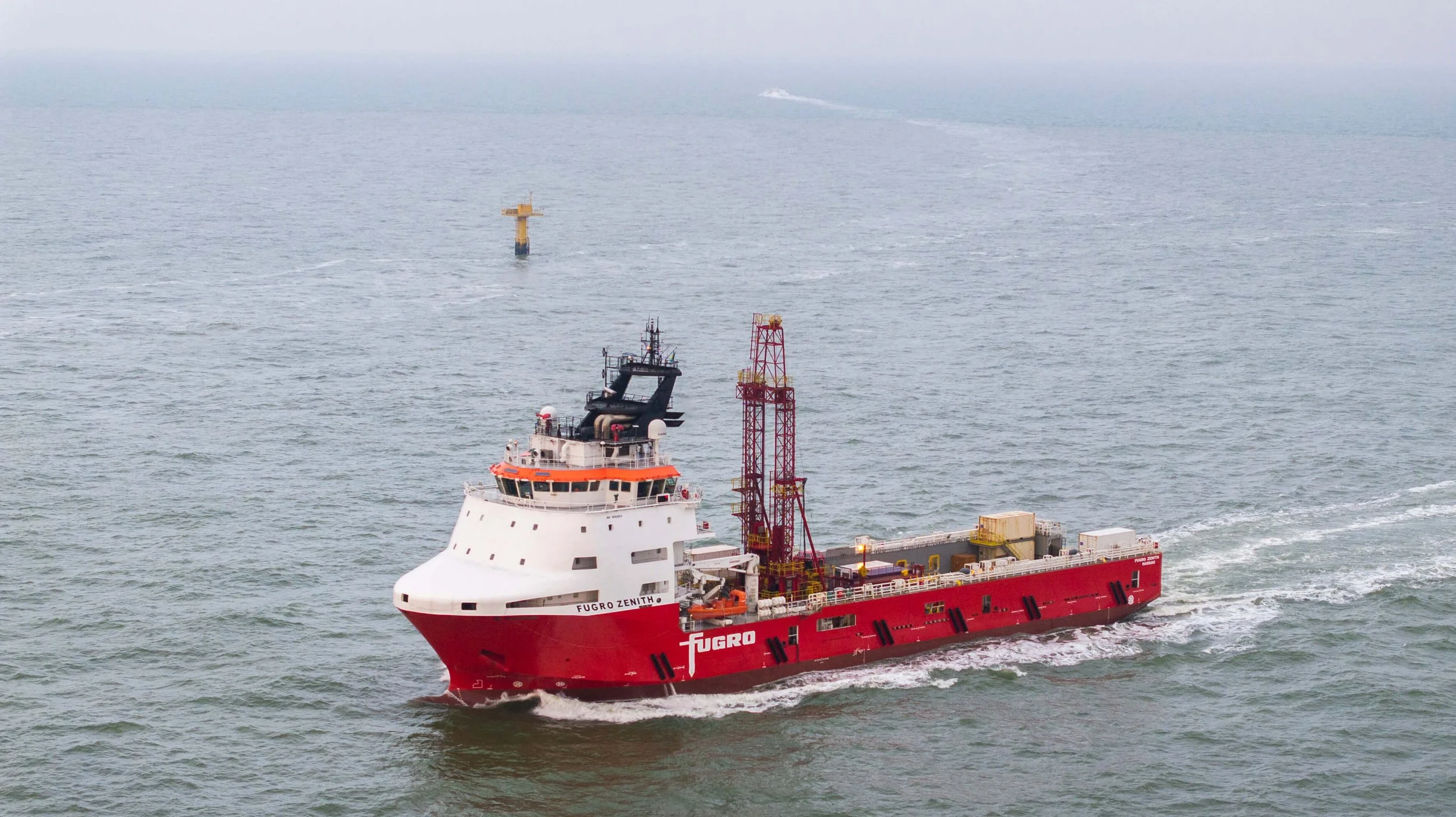 A red and white research or supply vessel named Fugro Zenitham navigating through open water, with a smaller yellow buoy or platform in the distance.