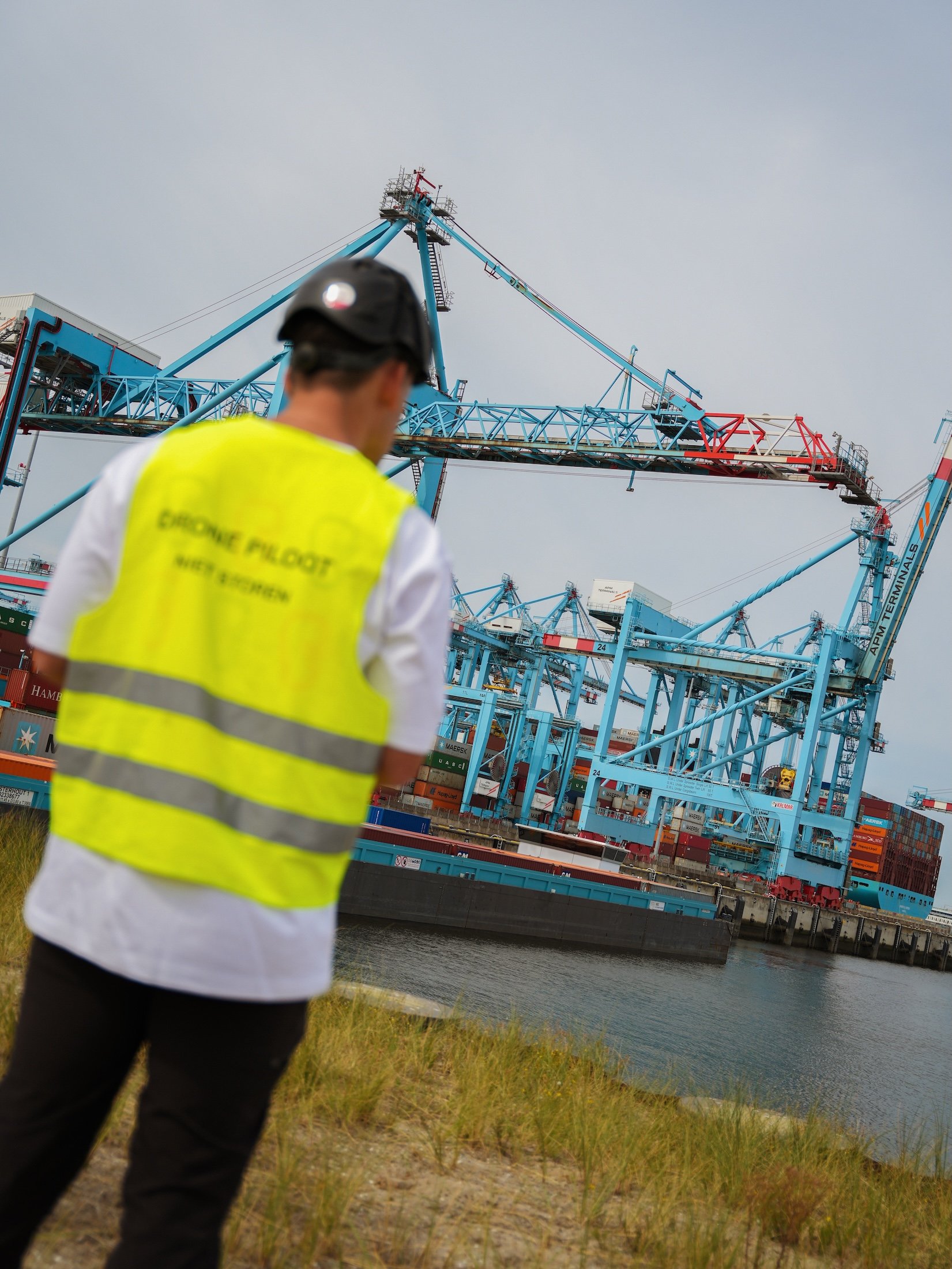 A person wearing a yellow safety vest and a black helmet standing near a body of water at a port or shipyard, with large blue cranes and cargo containers in the background.