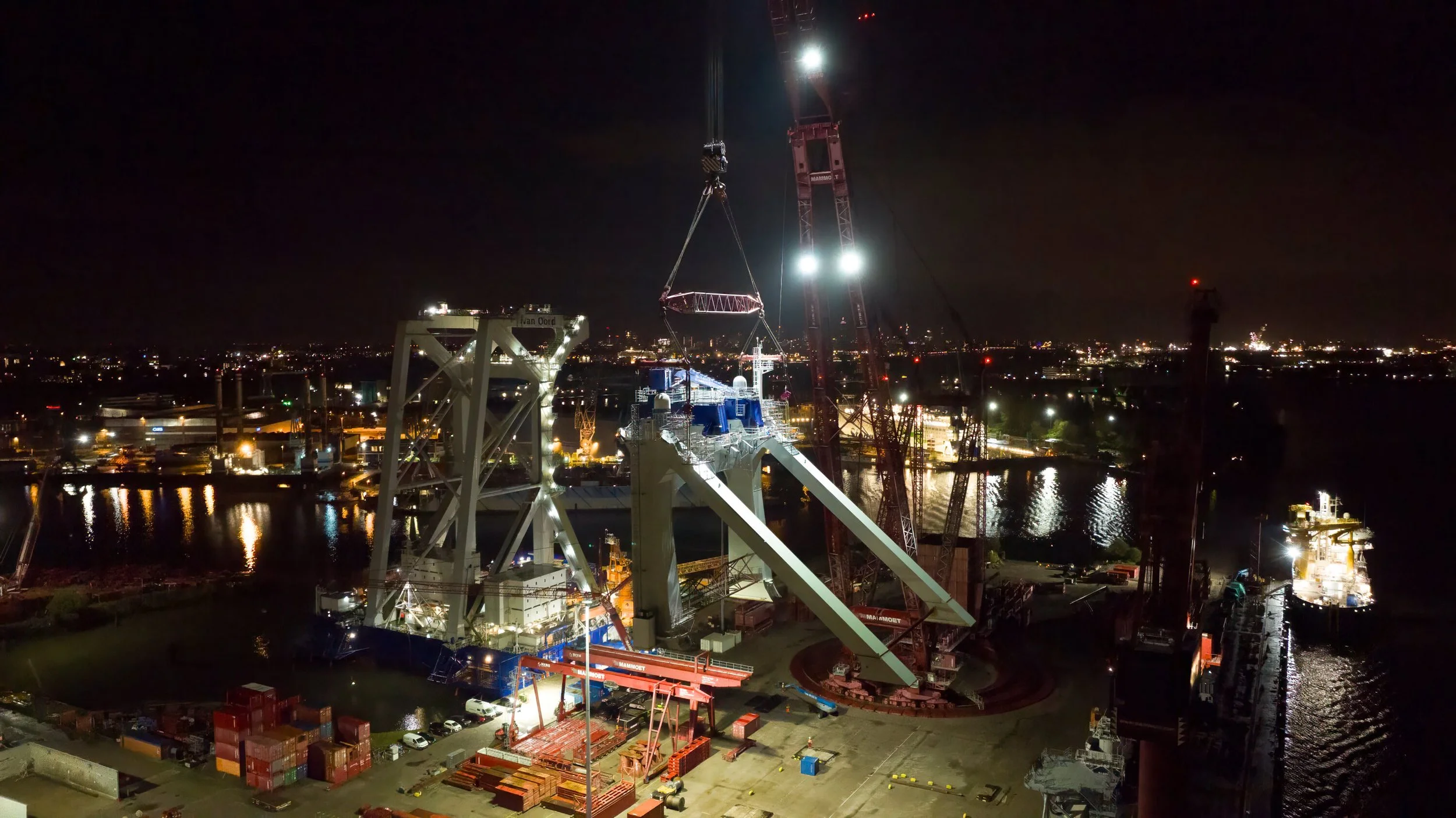 Nighttime scene of a large shipbuilding or dockyard with cranes installing a ship's superstructure on a vessel in a harbor, illuminated by industrial lights, with city lights reflected on the water in the background.